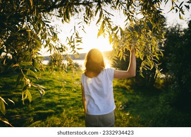 Young woman standing with her back in the rays of the sun at sunset on the banks of the river. Faith religion spirituality concept