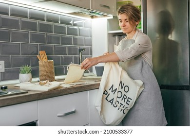 Young Woman Standing In The Kitchen Putting Reusable Cotton Produce Bags Into The Shopper Bag With Inscription NO MORE PLASTIC. Zero Waste Concept.