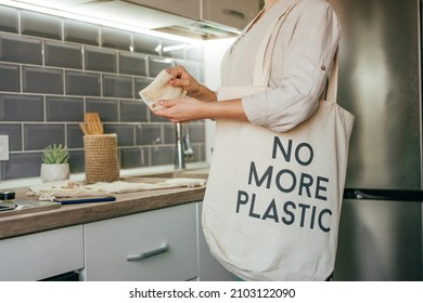 Young Woman Standing In The Kitchen Putting Reusable Cotton Produce Bags Into The Shopper Bag With Inscription NO MORE PLASTIC. Zero Waste Concept.