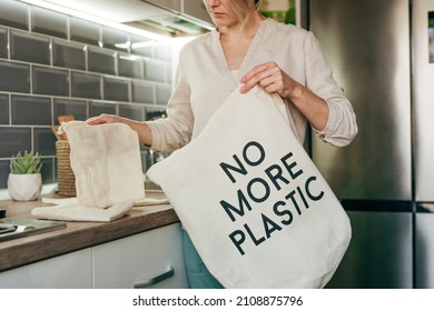 Young Woman Standing In The Kitchen Putting Reusable Cotton Produce Bags Into The Shopper Bag With Inscription NO MORE PLASTIC. Zero Waste Concept.