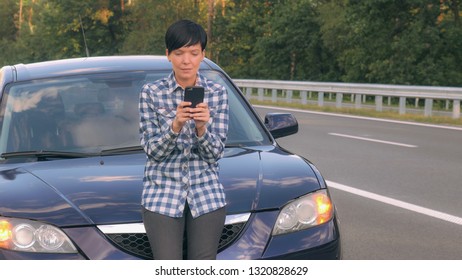 Young Woman Standing Near Broken Car Using Smartphone And Internet Search Phone Number Vehicle Service Or Local Garage. Brunette Wearing Casual Dress Travelling In Summer Season On The Driveway.