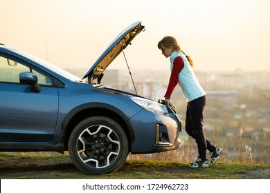 Young woman standing near broken down car with popped up hood having trouble with her vehicle. Female driver waiting for help beside malfunction auto.