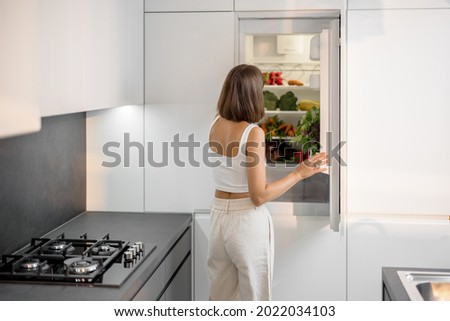 Young woman standing near the fridge full of fresh vegetables at modern kitchen. Healthy vegan eating concept