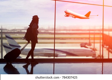 Young Woman Is Standing Near Window At The Airport And Watching Plane Before Departure. She Is Standing And Carrying Luggage.Travel Concept .