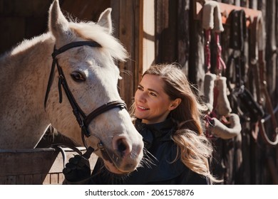 Young woman standing next to white Arabian horse resting in wooden stables box, sun shines on them, closeup detail