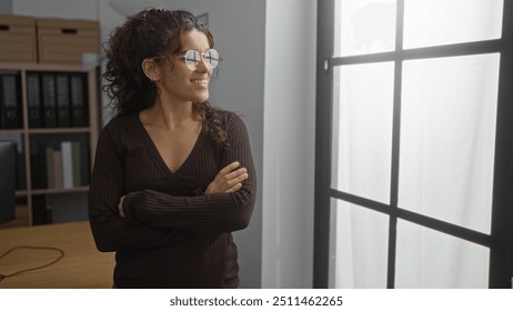 Young woman standing in an office with arms crossed looking through a window, wearing glasses and a dark shirt, in a well-lit workplace setting. - Powered by Shutterstock