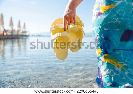 Young woman standing on beach. Low angle shot of woman in blue dress holding yellow sandals in hand and standing against seascape. Cropped shot. Leisure, vacation concept