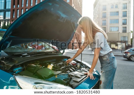 Young woman standing on the city street while examining her broken down car with open hood, Selective focus, Horizontal shot
