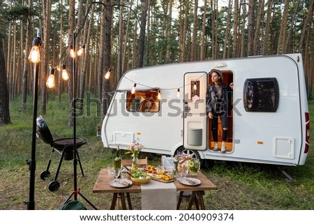 Young woman standing in open door of house on wheels among pinetrees and served table in front