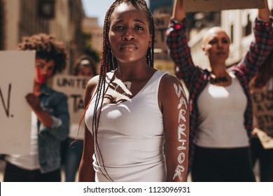 Young Woman Standing Outdoors With Group Of Protesters At Back. Woman With Word Warrior Written On Her Arm.
