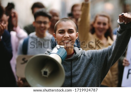Young woman standing outdoors with group of demonstrator in background. Woman protesting with a megaphone outdoors on road.