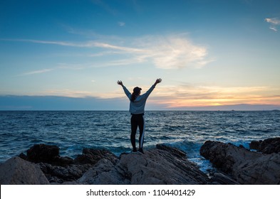 Young Woman Standing Outdoors At Sunset Close Adriatic Sea In Croatia. Beautiful Nature And Landscape On Dusk Evening At The Ocean In Dalmatia. Girl Holding Arms Up In Air. Calm, Peaceful And Happy.