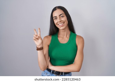 Young woman standing over isolated background smiling with happy face winking at the camera doing victory sign with fingers. number two. 