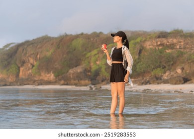 A young woman standing in shallow water at the beach, holding a drink. She wears a black crop top and a black skirt, with a light shirt and a cap. The background features a rocky coastline - Powered by Shutterstock