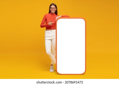Young woman standing and showing with finger blank screen of huge phone, mockup for your app, isolated on yellow 