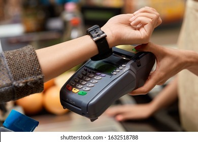 Young woman standing at the supermarket at cashier checkout counter paying for purchases contactless with smartwatch applying on reader doing daily shopping close-up - Powered by Shutterstock