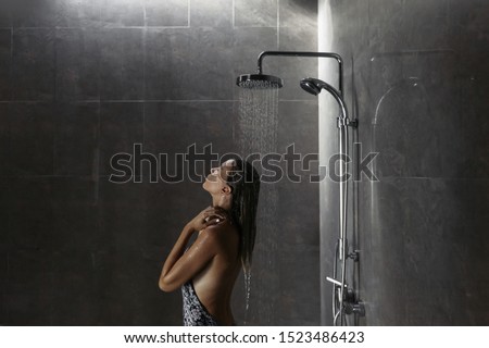 Young woman standing under shower and washing hair back to the camera over black ceramic tiles in modern luxury dark bathroom interior.
