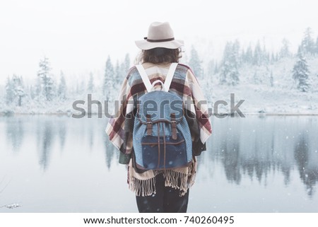 Young woman standing in the wild in front of amazing natural lake view. Wearing hat, poncho and backpack. Winter is coming, first snowfall. Wanderlust and boho style