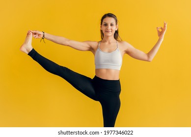 Young Woman Standing In Yoga Asana Utthita Hasta Padangusthasana, Extended Hand To Big Toe Yoga Pose , Studio Shot, Yellow Background