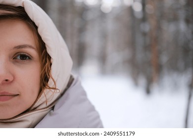 A young woman stands against a background of fir trees under a snowy forest in winter. A girl in a gray down jacket with a hood stands and looks at the camera. Close-up. - Powered by Shutterstock