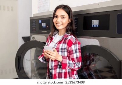 A young woman stands by a washing machine in a laundromat, smiling as she checks her smartphone. The environment is bright and contemporary, making laundry day pleasant. - Powered by Shutterstock