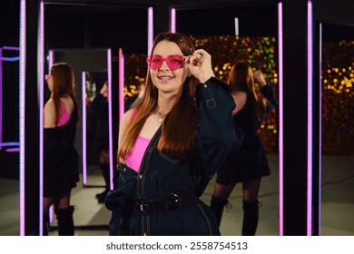 A young woman stands confidently in a stylish outfit, holding pink sunglasses. The vibrant room is filled with mirrors and colorful lights, creating a lively and fashionable atmosphere. - Powered by Shutterstock