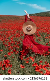 Young Woman Stands With Her Back In A Long Red Dress And Hat, Posing On A Large Field Of Red Poppies