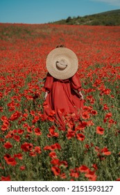 Young Woman Stands With Her Back In A Long Red Dress And Hat, Posing On A Large Field Of Red Poppies