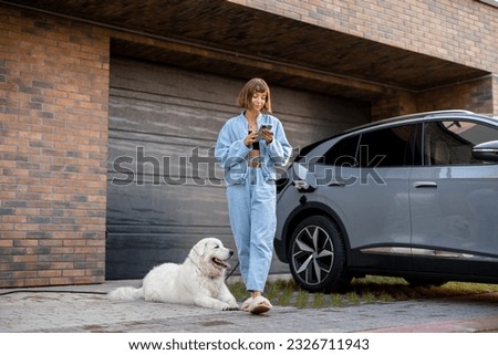 Young woman stands with her dog and using smart phone while charging her electric car near garage of her house. Concept of modern lifestyle and sustainability
