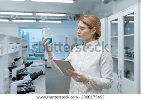 A young woman stands in the laboratory in a white coat and protective glasses, holds a flask with a blue substance and a tablet in her hands, studies the contents and composition of the sample.