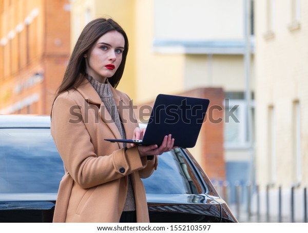 a young woman stands with a laptop\
near her car. She is serious and looking for something on the\
Internet. concept of car insurance and roadside\
assistance