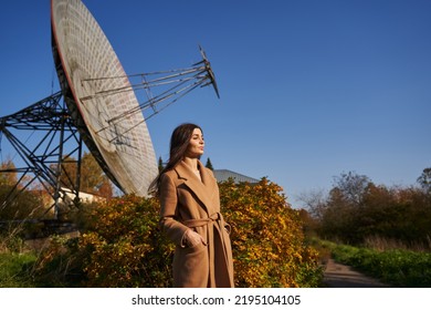 a young woman stands near a huge astronomical radar in a brown coat, surrounded by autumn nature and a small building and a clear blue sky - Powered by Shutterstock