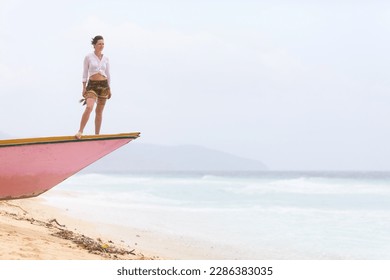 A young woman stands on the bow of a wooden boat and looks out into the ocean