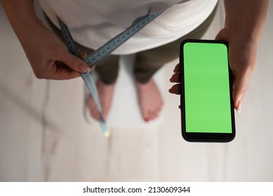Young woman stands on white electronic scale with measure tape. Girl holds mobile phone with app controls, loses weight for sports, diet. Measurement in kg. Smartphone device. Chroma key green screen.