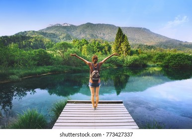 Young Woman Stands On A Wooden Bridge With Raised Arms Up On The Nature Background. Travel, Freedom, Lifestyle Concept. Slovenia, Europe.