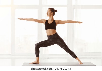 A young woman stands on a yoga mat, arms extended in Warrior II pose, exuding tranquility and focus in a sunlit, spacious studio room - Powered by Shutterstock