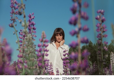 Young woman stands in white shirt in field of purple lupins against blue sky