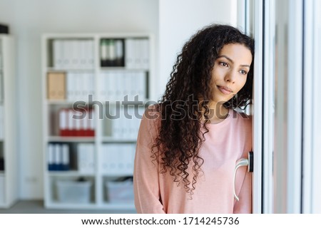 Young woman staring longingly through a window with a sad faraway expression as she leans against a wall in an office