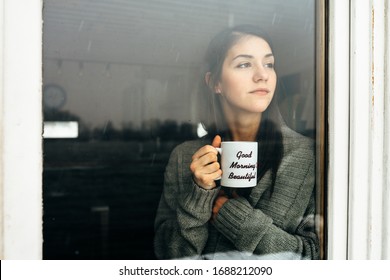 Young Woman Staying At Home Drinking Coffee/tea,looking Trough The Window.Starting The Day,morning Ritual.Quaratine Self Isolation Concept.Worried Woman Indoors Activity.Mental Challenge.Bad Mood