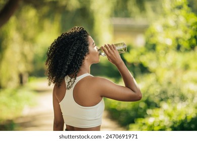 A young woman stays hydrated by drinking water from a bottle while exercising in a green, sunlit park on a beautiful morning. - Powered by Shutterstock