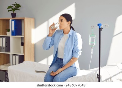 Young woman with sterile needle in arm sitting on medical bed by pole with IV drip bottle, drinking glass of water and receiving vitamin therapy infusion to boost immune system and suppress infection - Powered by Shutterstock