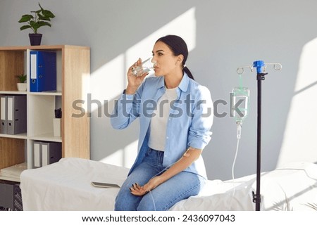 Young woman with sterile needle in arm sitting on medical bed by pole with IV drip bottle, drinking glass of water and receiving vitamin therapy infusion to boost immune system and suppress infection