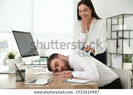 Young woman sticking paper fish to colleague's back while he sleeping in office. Funny joke