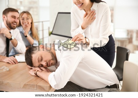 Young woman sticking paper fish to colleague's back while he sleeping in office, closeup. Funny joke