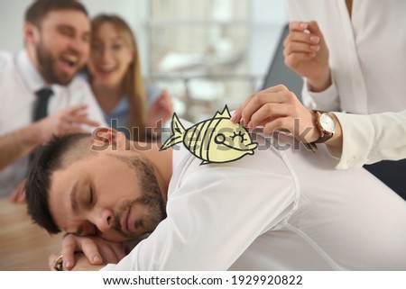Young woman sticking paper fish to colleague's back while he sleeping in office, closeup. Funny joke