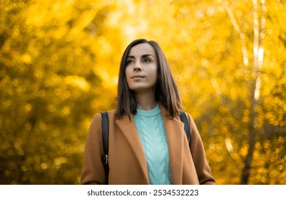 Young woman with straight brown hair stands outdoors in sunlit autumn park. She wears beige coat and blue sweater, looking thoughtfully into the distance, with vibrant yellow foliage behind her - Powered by Shutterstock