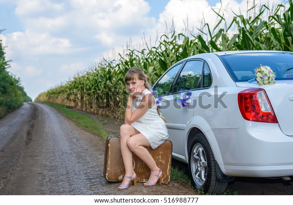 Young woman stranded at the roadside sitting on a\
battered old suitcase alongside a car decorated with flowers in an\
field of young corn