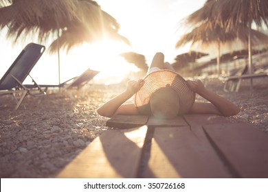 Young Woman In Straw Hat Lying On A Tropical Beach,enjoying Sand And Sunset.Laying In The Shade Of Palm Tree Parasols.Summer Vacation Concept.