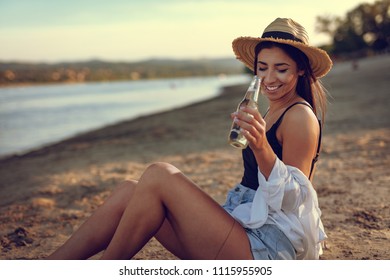 Young woman in straw hat relaxing at sunset time on the river bank. She is sitting on the river send and drinking beer. 