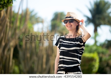 Young woman in straw hat, sunglasses and summer dress walking on tropical resort street, happy smiling